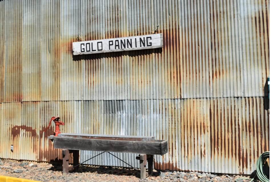 a metal wall with a Gold Panning sign and a wooden bench below it.