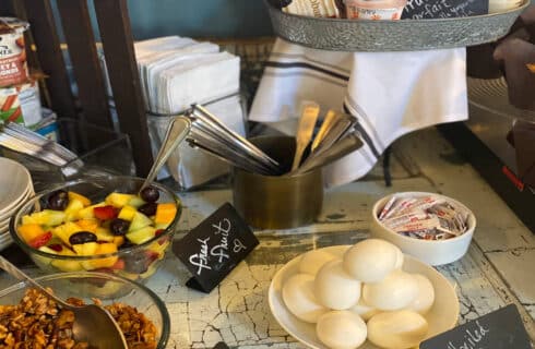 a table with breakfast foods including hard boiled eggs, granola, fresh fruit, oatmeal with bowls, spoons and napkins.