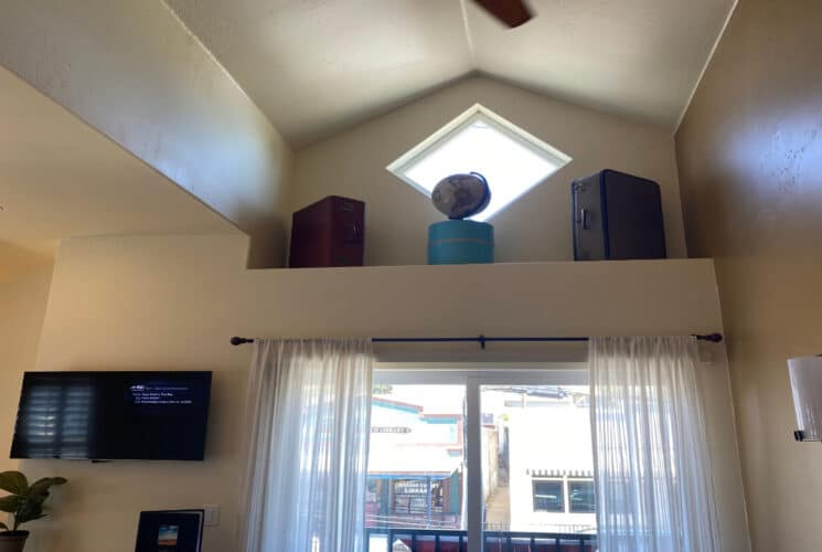 a room with tall windows with sheer curtains leading outside, a high shelf with a diamond shaped window and vintage suitcases and a globe on the shelf, and a flat screen tv on the wall next to the window.