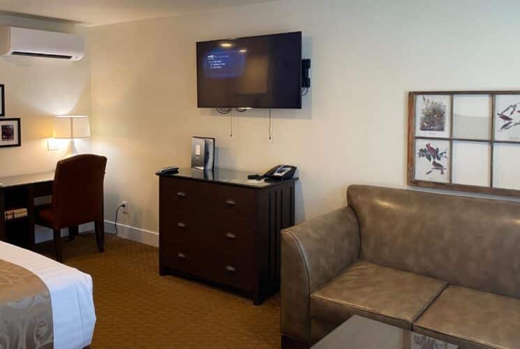 a bedroom with a bed on one wall, a sofa, dresser and flat screen tv on another wall, and a writing desk with lamp and chair in the corner.