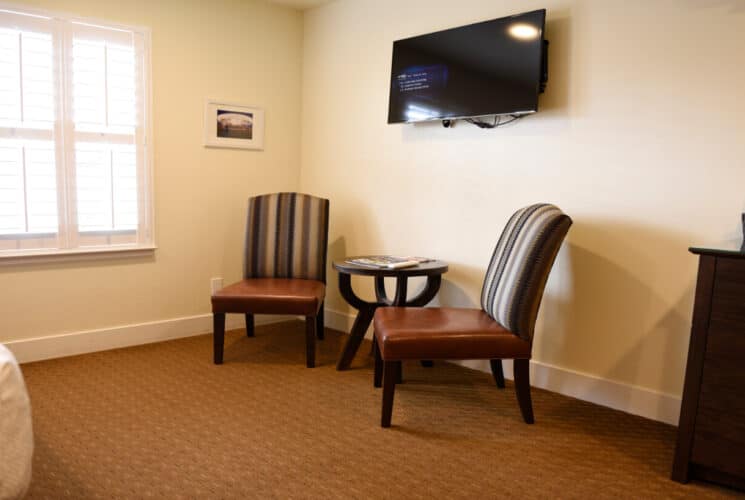 the corner of a bedroom with 2 chairs with a small table in between them, a flat screen TV above them, and a dresser.