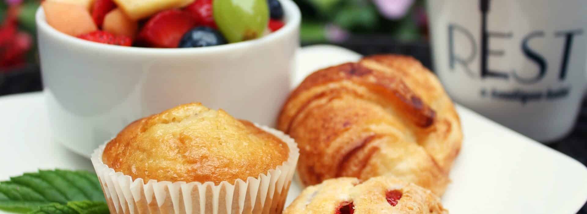 muffins and pastries on a plate with a bowl of fruit, and a mug that says REST with a key