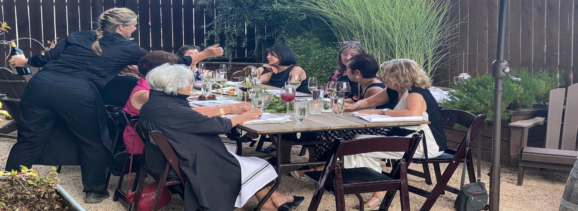 an outdoor table with several people enjoying an outdoor meal with glasses of wine