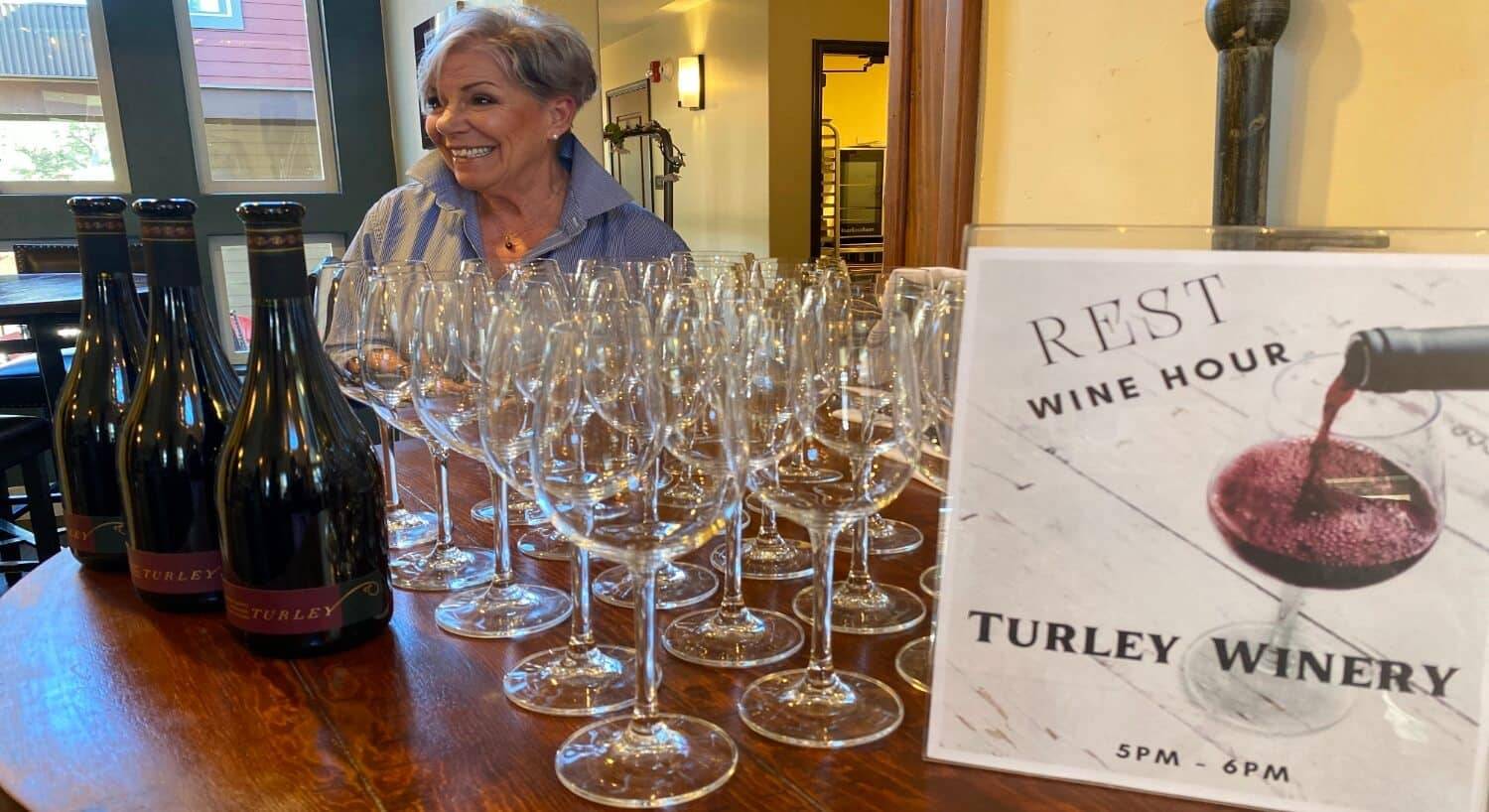 a table with bottles of wine and lots of wine glasses, and a sign that says Rest Wine Hour and Turley Winery, and a smiling woman