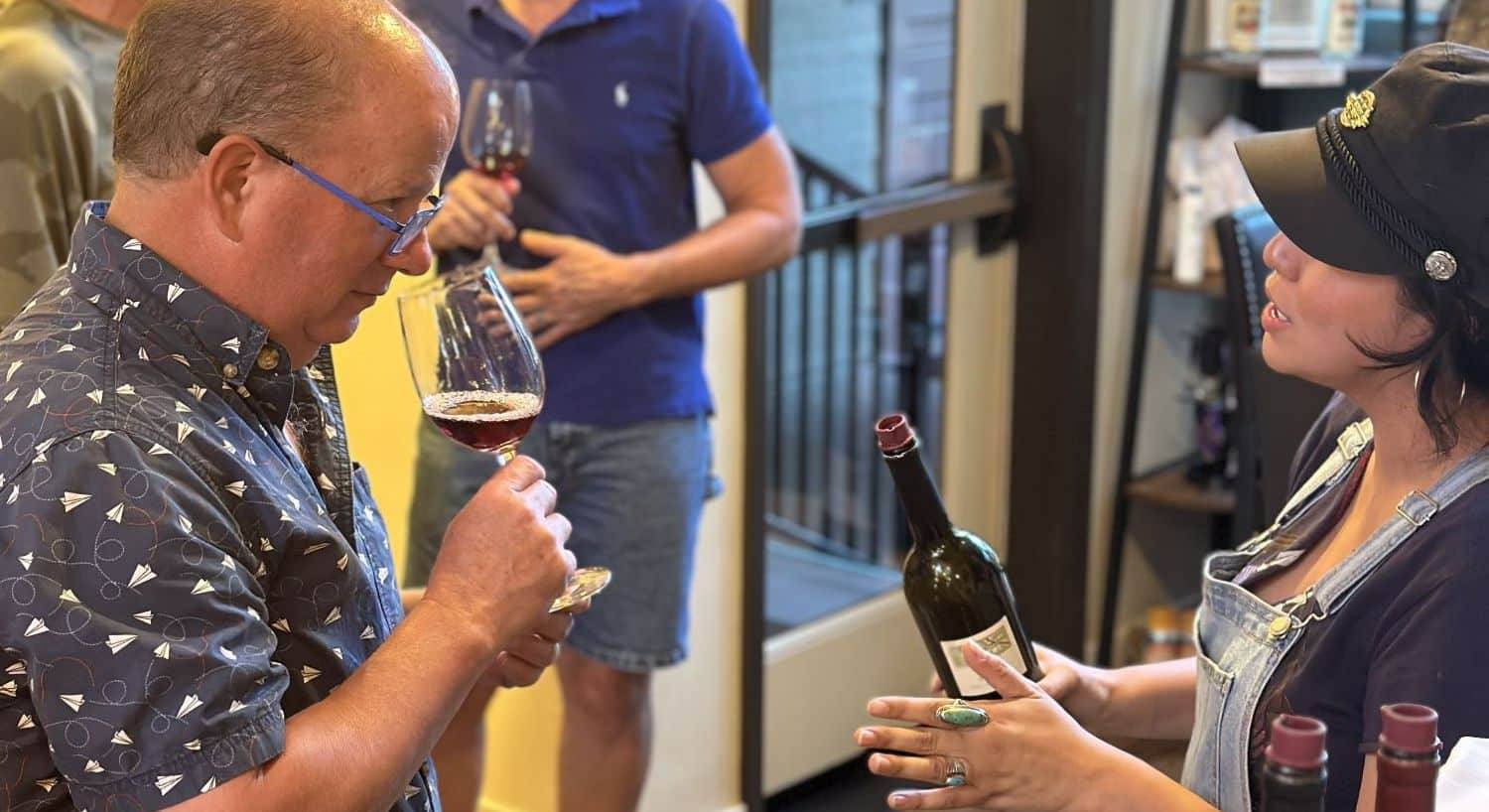 a woman in a hat with a wine bottle in hand speaking with a man smelling the wine