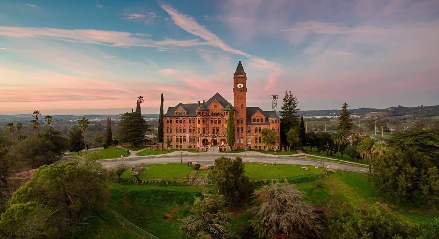 a red brick building with a clock tower in the midst of green grass, trees, and a colorful sky