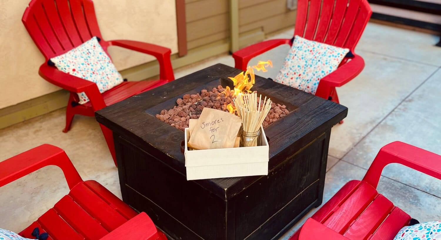 an outdoor square firepit with a s'mores kit on one edge, and 4 red chairs around it.