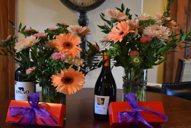 A bouquet of colorful orange flowers with a bottle of champagne and red boxes of chocolates with purple ribbon