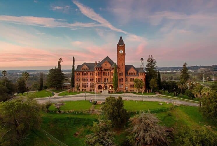 a red brick castle with a tall clock tower amidst green grass, bushes and trees, and a blue and pink sky