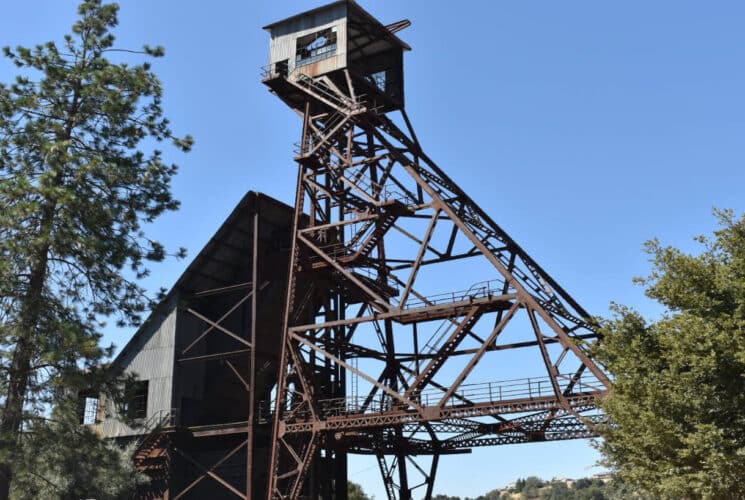 a mine shaft and tower next with a pine tree and green bushes on either side, and blue skies above